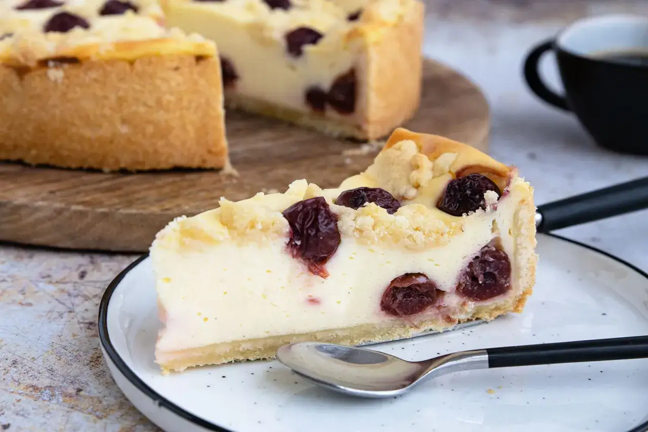 Ein saftiges St&uuml;ck Kirschkuchen, ein k&ouml;stlicher Kuchen ohne Ei, mit Streuseln und vielen Kirschen, serviert auf einem Teller mit einer Kaffeetasse im Hintergrund.